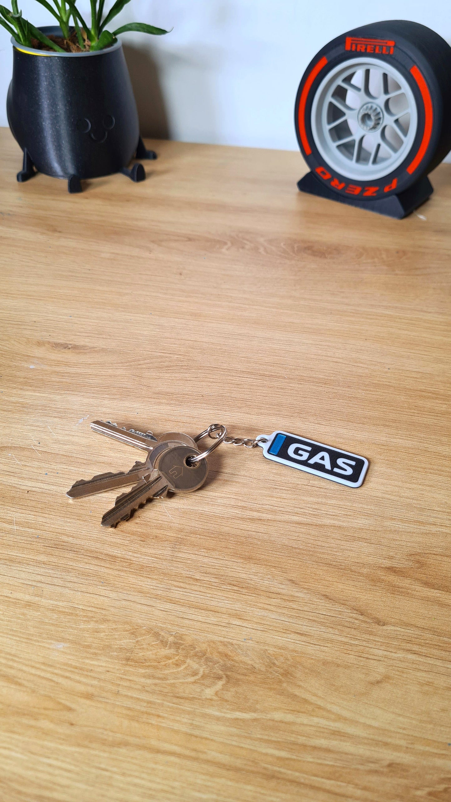 Personalized F1-inspired keychain featuring the tag "GAS" on a wooden table. Includes standard keys attached, with decoration including a cartoon plant pot and a Pirelli tire clock in the background.