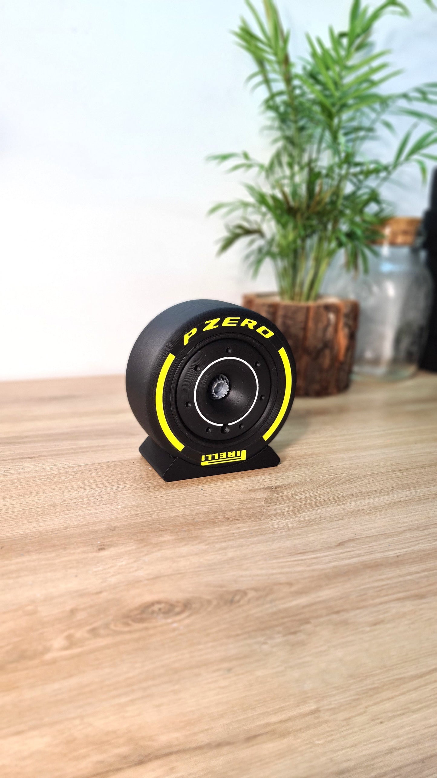 Image shows a customizable Formula 1 tyre rim cover with a nut, displayed on a wooden surface. The rim cover is black with red detailing and the Pirelli P Zero logo. A potted plant in the background adds contrast.