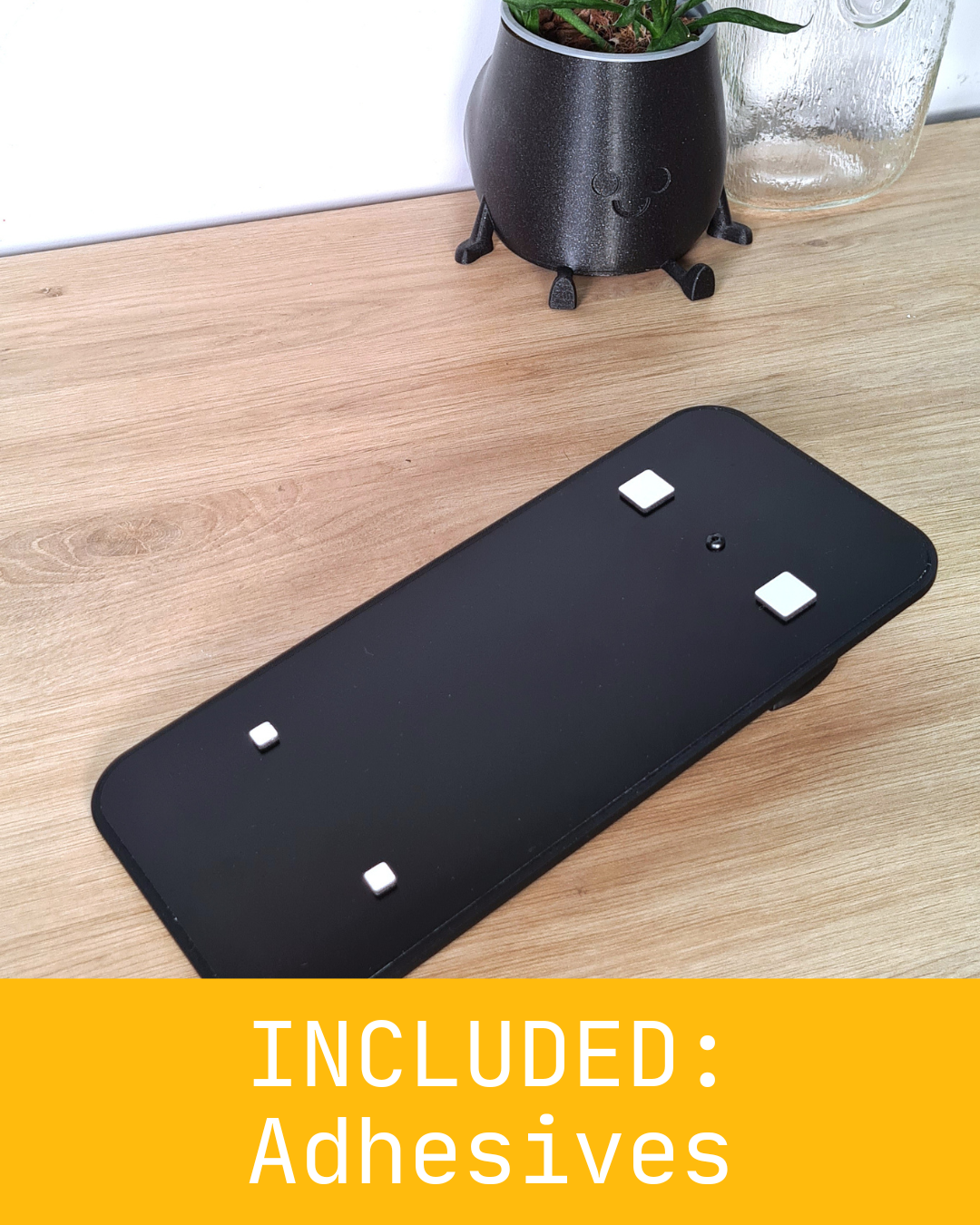 Alt Text: A rectangular black object labeled as "INCLUDED: Adhesives", equipped with small white adhesives on its surface. Placed on a wooden table near a smiling pot and a glass jar.