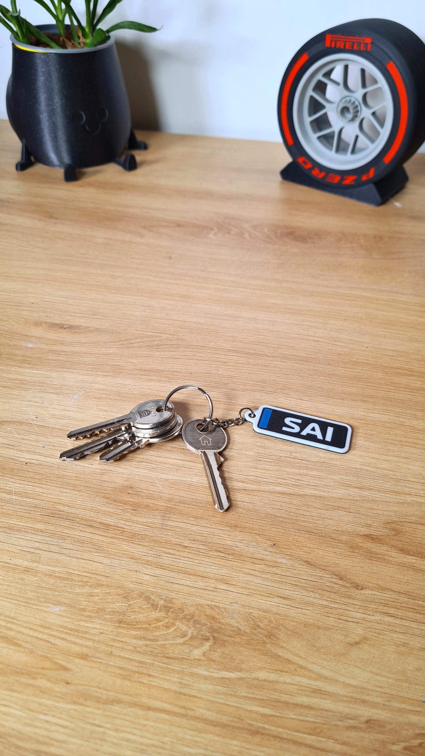 Personalized Williams F1 car keychain labeled "SAI" on a wooden desk, next to keys and a decorative plant pot, with a mini Pirelli tire model in the background. Ideal for racing fans.