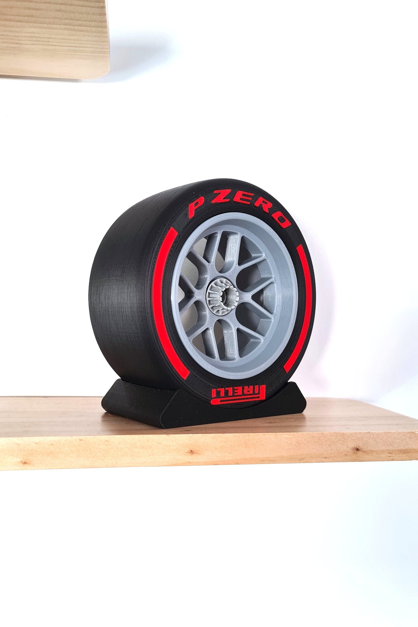 A replica of an F1 tire and rim, "P Zero" prominently displayed in red with a silver rim, mounted on a black stand, on a light wooden shelf against a white background.