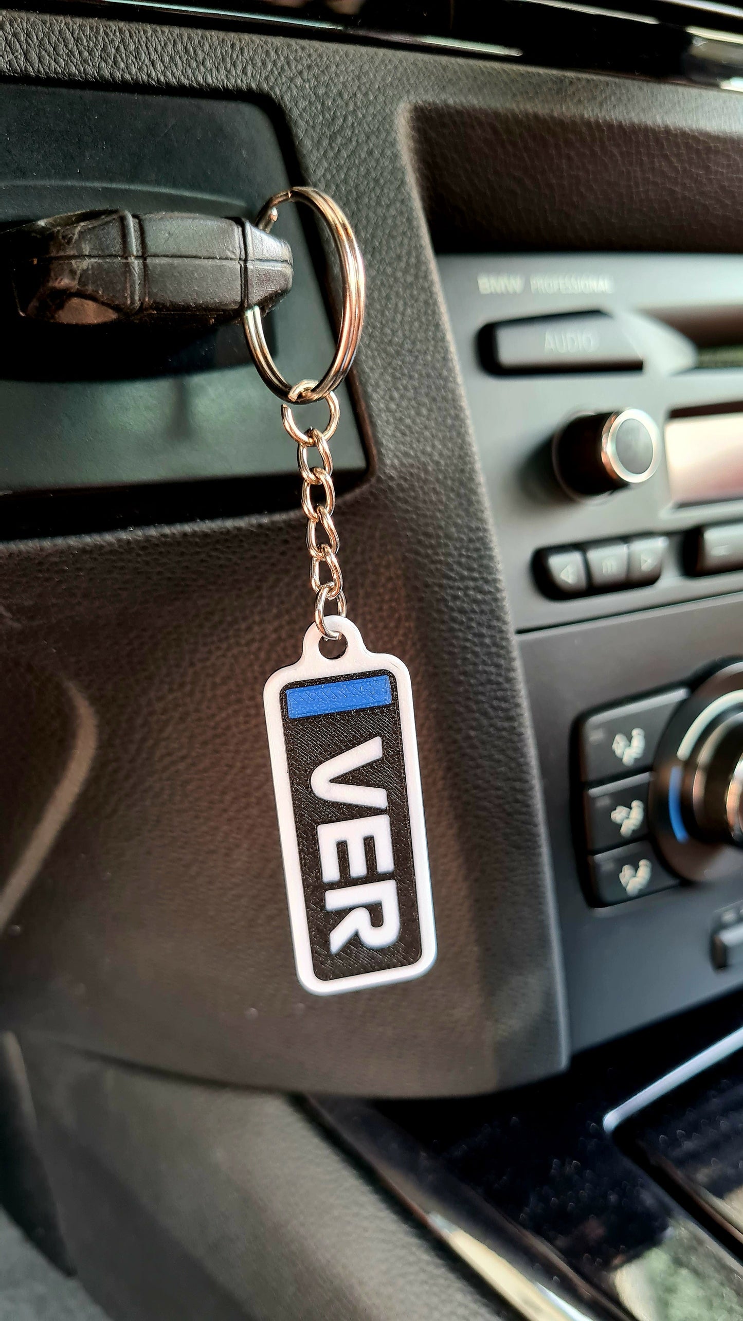 A customizable Formula 1-themed keychain hanging in a car, displaying "VER" in bold white letters with a blue stripe, set against a black car dashboard background. Ideal for motorsport fans.