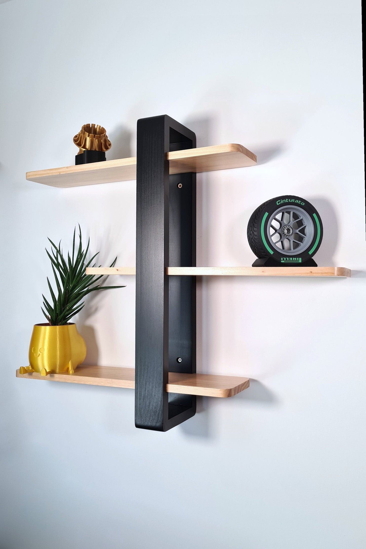 Decorative wall shelf displaying objects including a replica of an F1 tyre and rim, a yellow pot with a green plant, and a gold elephant figurine. The shelf features a black support and light wood.