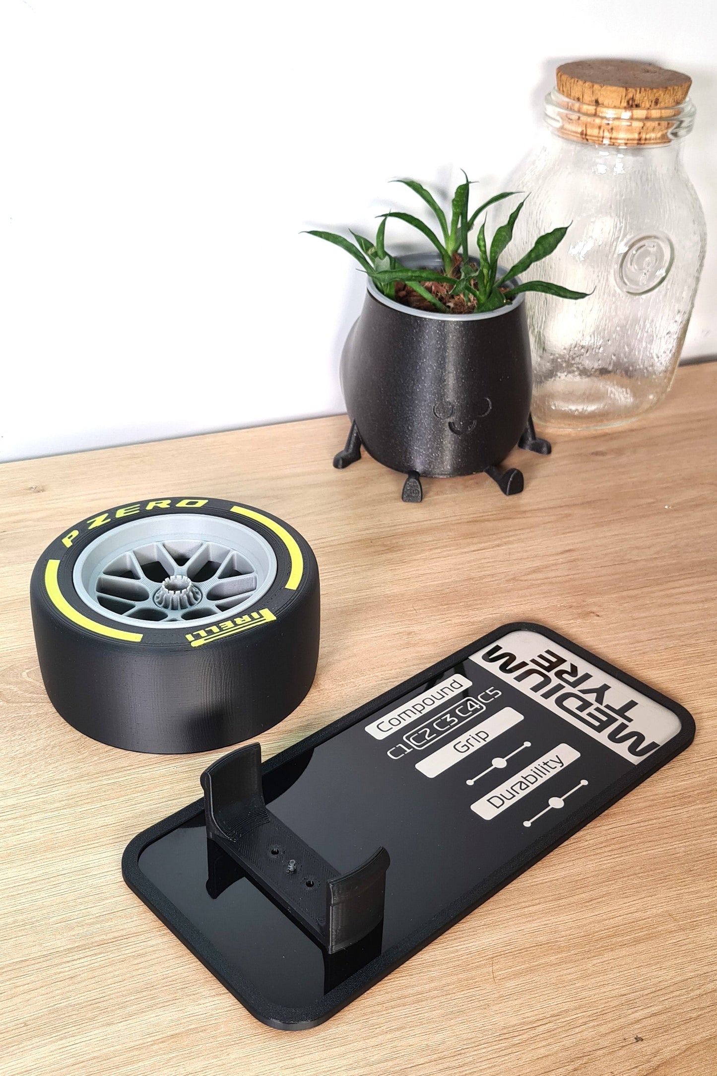 Formula 1 tire and rim replica displayed next to a specs panel showing compound details. Ideal gift for racing enthusiasts. Decor includes a happy-face pot with a plant and a glass jar on a wooden surface.
