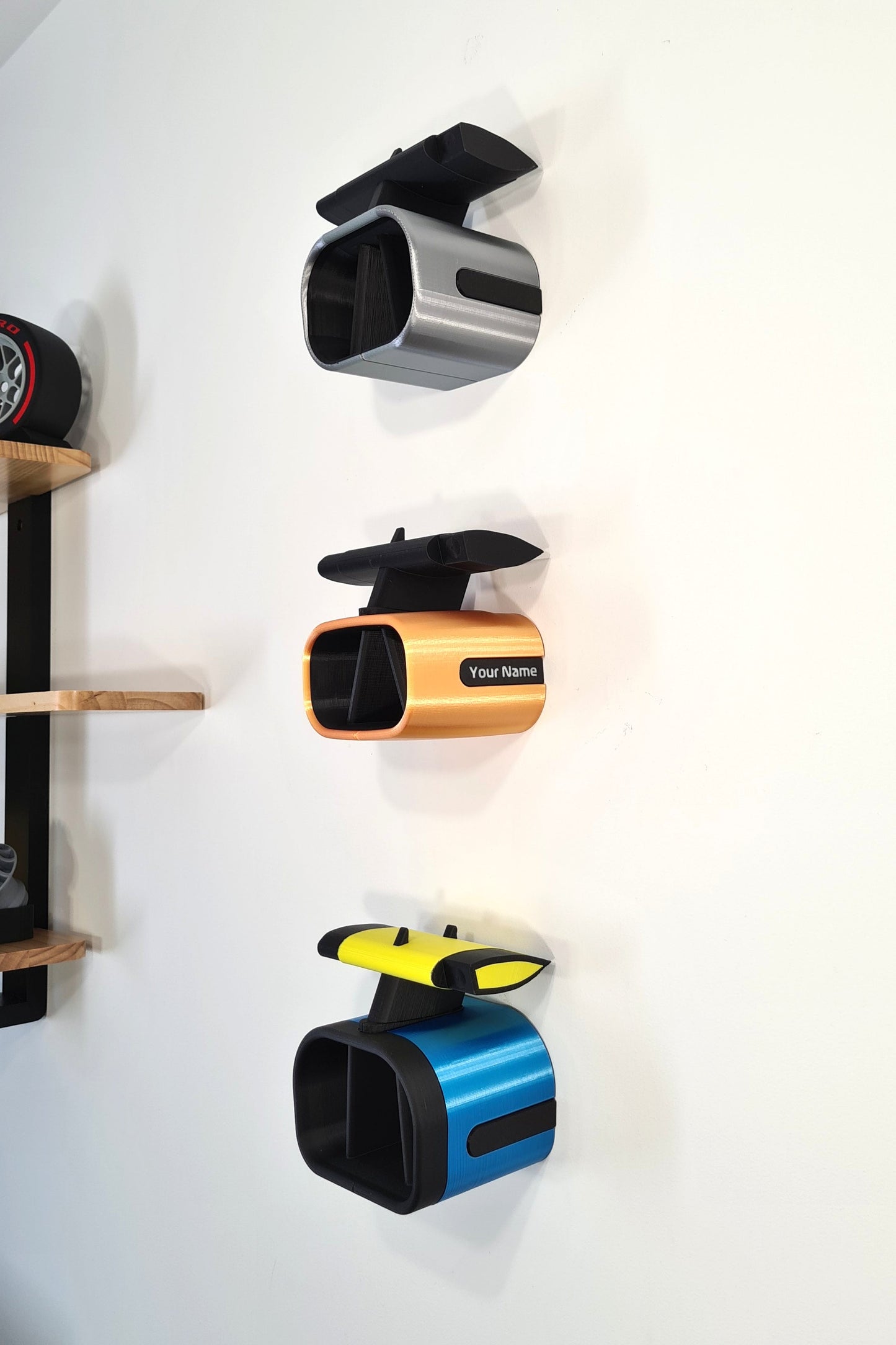 Wall-mounted replica Alpine Formula 1 airboxes in various designs, including one customizable with a name, displayed on a white wall beside wooden shelves. Ideal for motorsport enthusiasts.