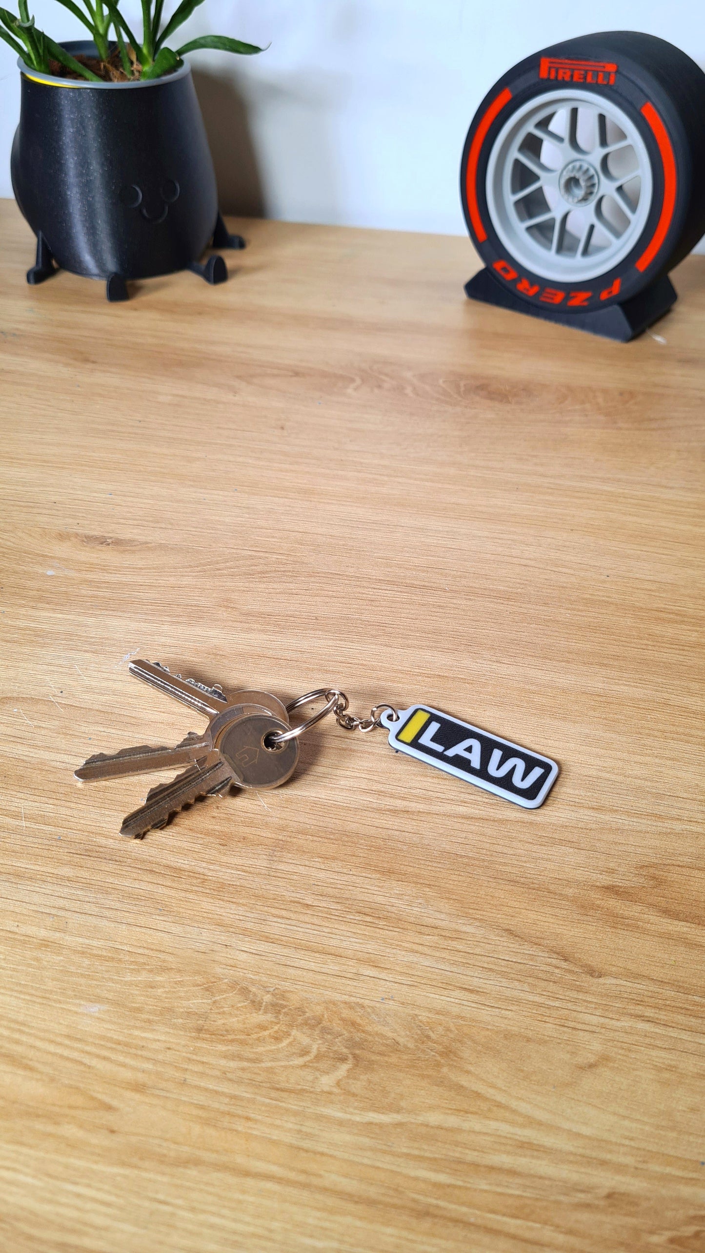 A personalized keychain with the acronym "LAW" in yellow, black, and white on a desk, attached to several keys, near a black plant pot and a Pirelli tire clock.