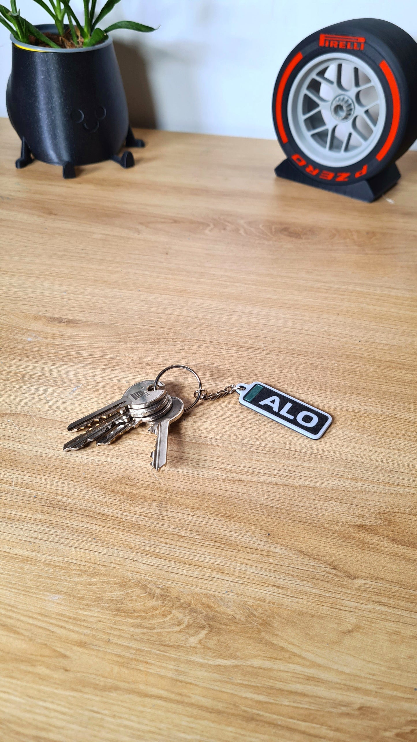 A keychain with the text "ALO," attached to a set of keys on a wooden table, next to a plant in a black pot and a Pirelli tire clock. Ideal for F1 fans who prefer personalized merchandise.