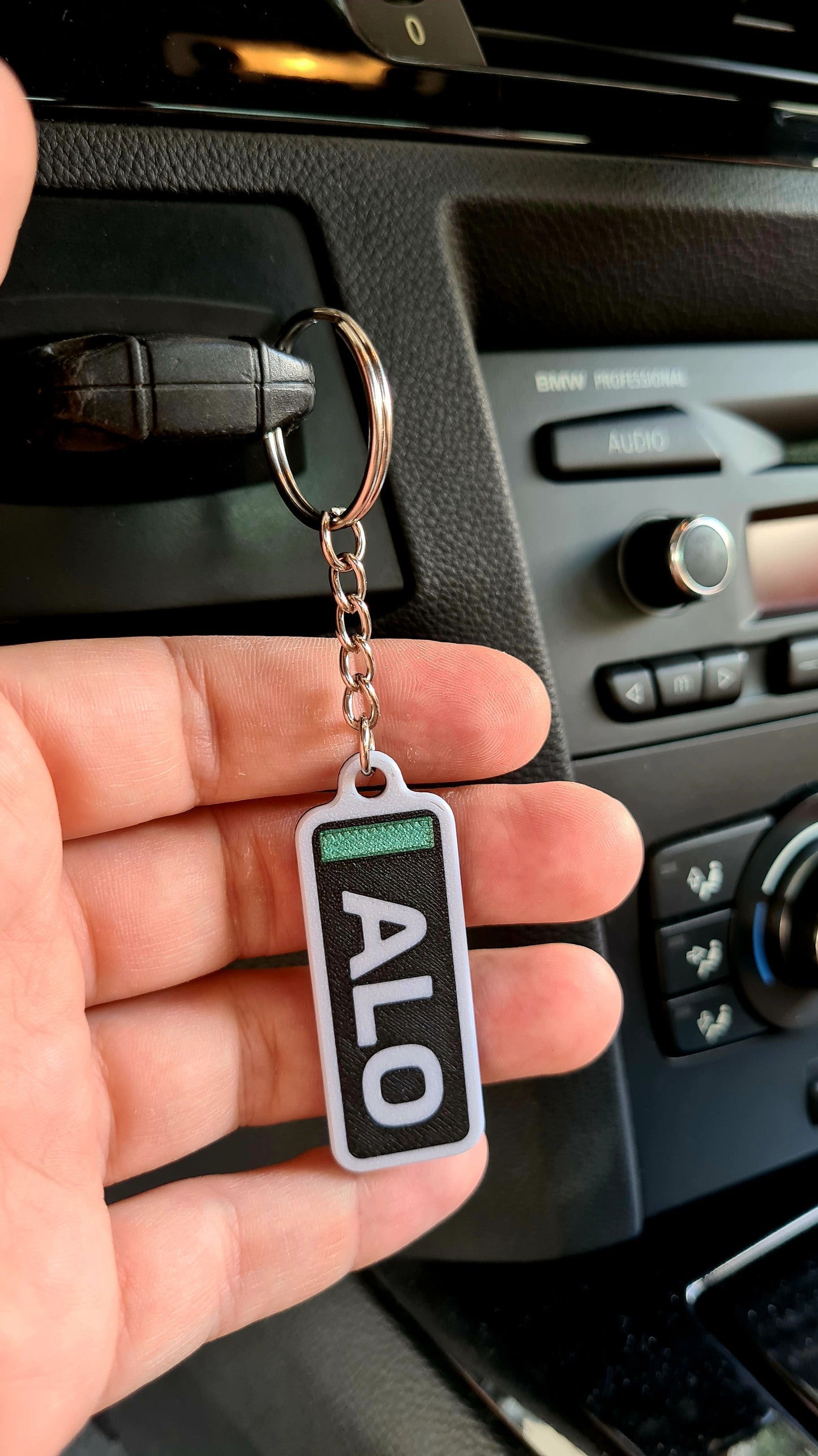 A personalized keychain with the letters "ALO" on a wooden table, alongside house keys, beside a decorative pot and a miniature tire model. Ideal for Formula 1 fans to customize with their favorite driver's initials.