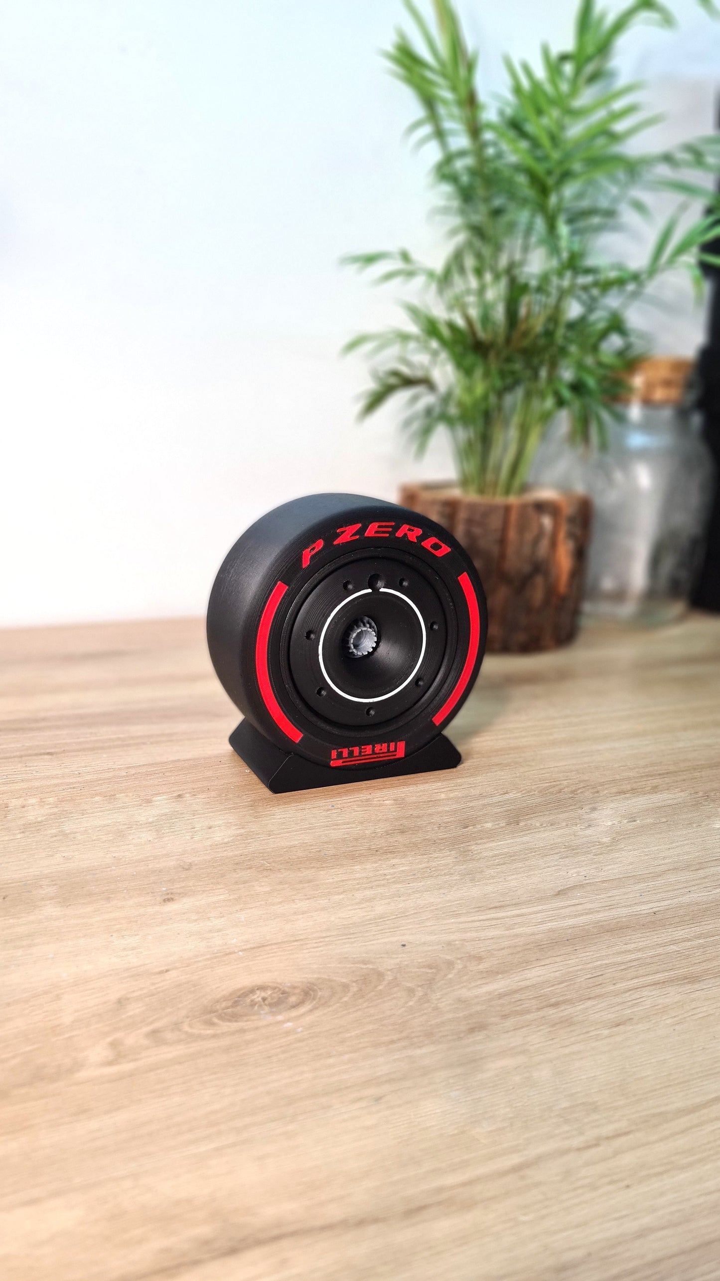 A customizable Formula 1 tyre rim cover and nut, Pirelli P Zero version, displayed on a wooden surface with a blurred plant in the background. This mini replica features a sleek black and red design.
