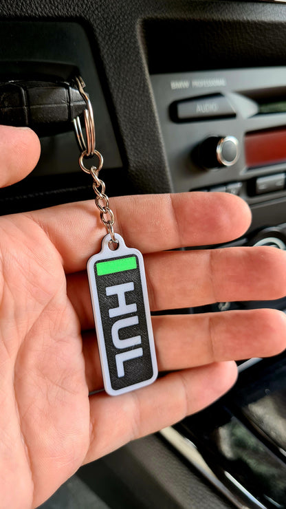 Personalized F1-themed keychain with the abbreviation "HUL" displayed in black and white, attached to a set of metal keys on a wooden table, featuring a Pirelli tire desk clock and a black planter in the background.