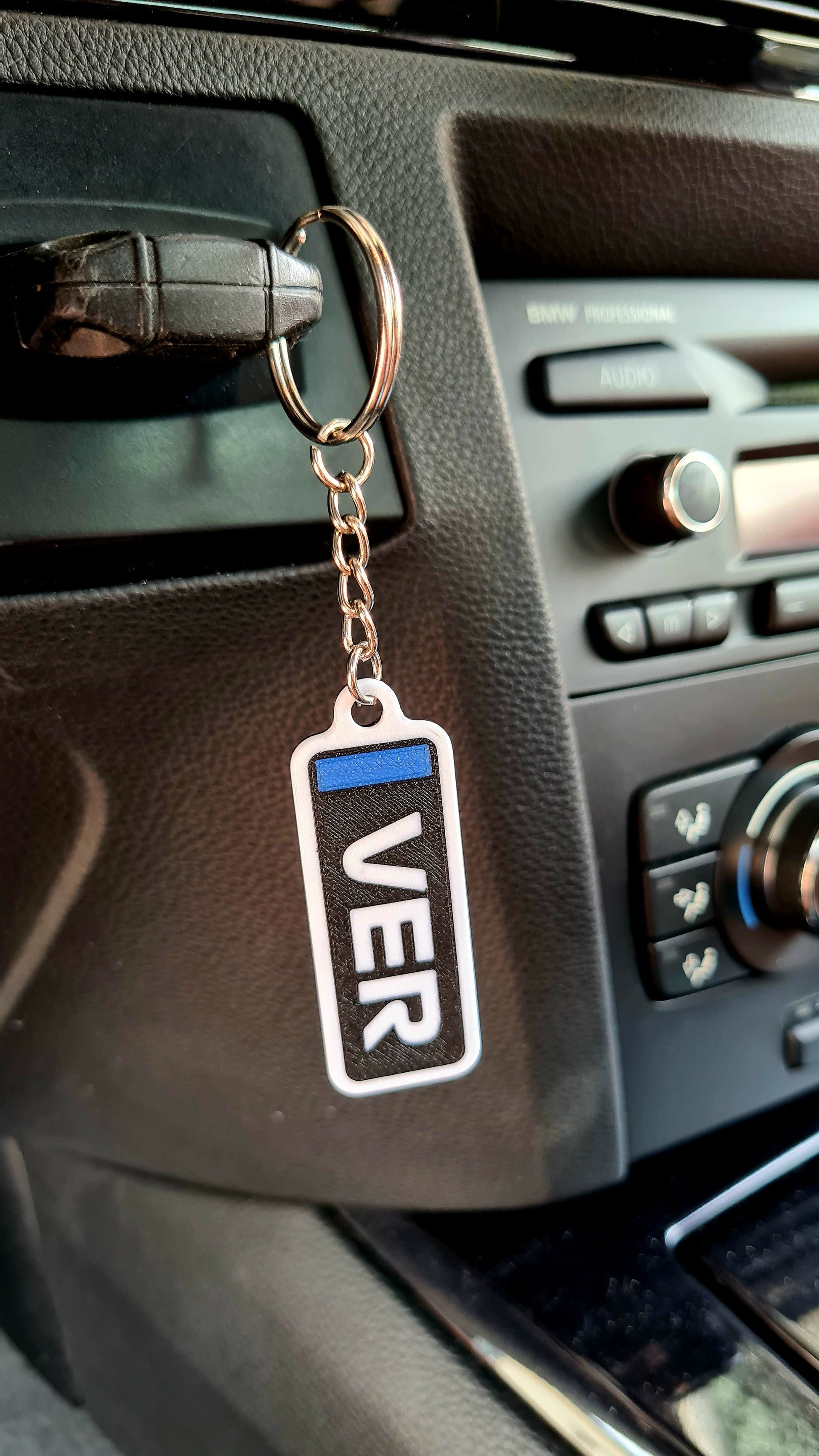 A customizable Formula 1-themed keychain hanging in a car, displaying "VER" in bold white letters with a blue stripe, set against a black car dashboard background. Ideal for motorsport fans.