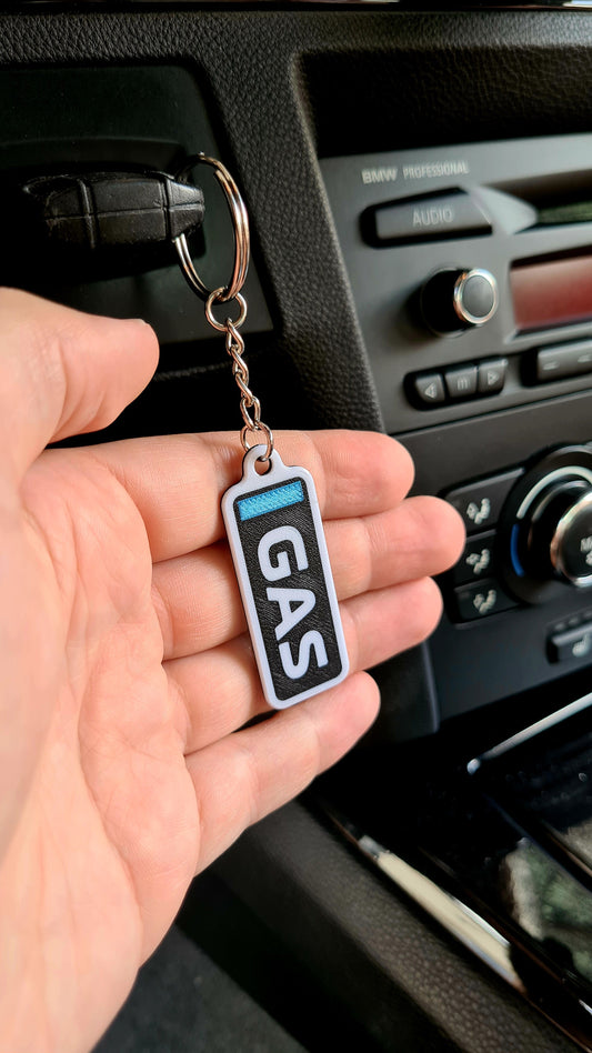 Personalized F1-inspired keychain with "GAS" text in blue and white on a wooden table, next to keys, a happy face plant pot, and a Pirelli tire clock. Ideal for racing fans and car enthusiasts. Highly detailed and vibrant colors.