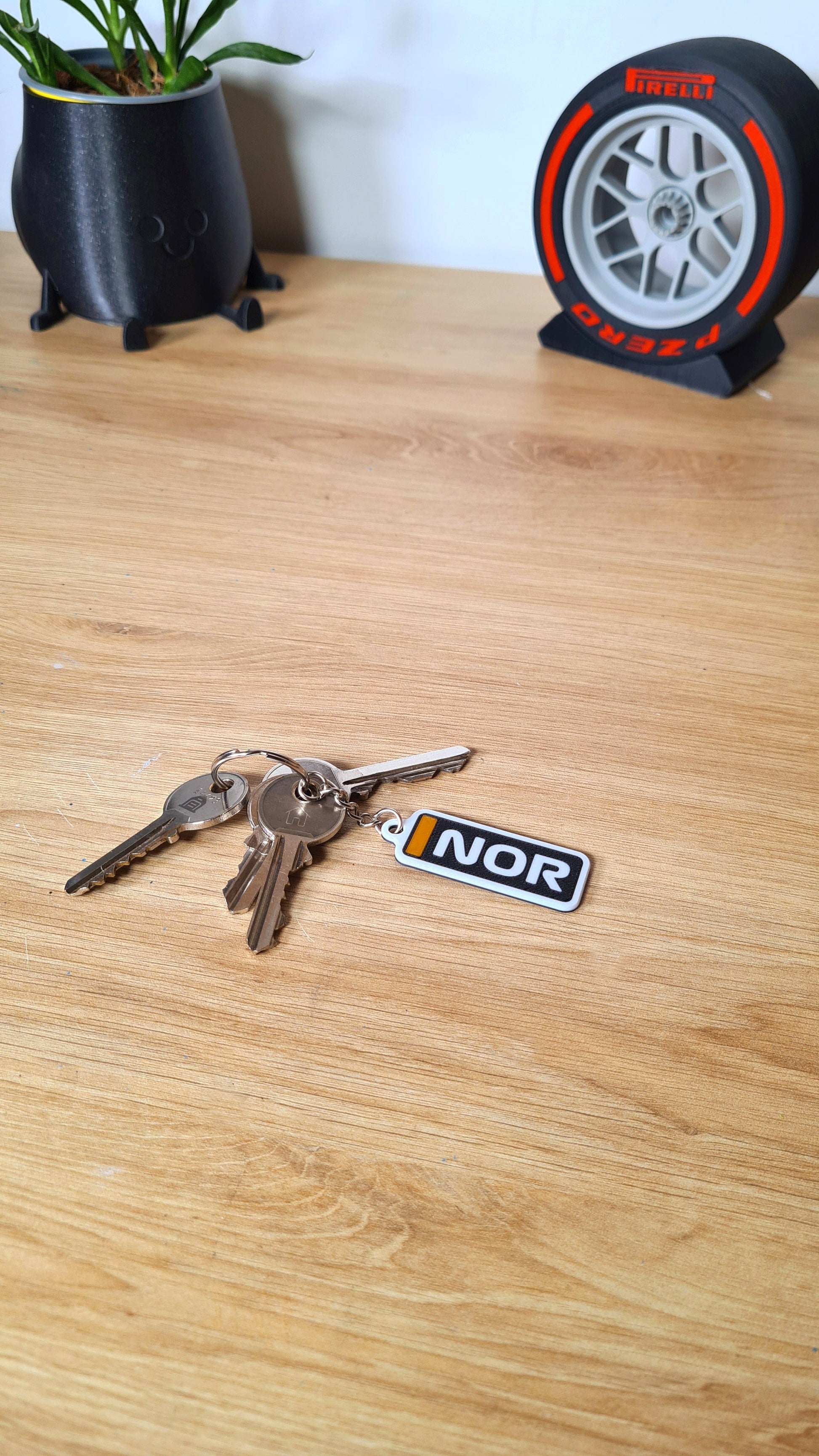 Personalized McLaren F1 car keychain with custom "NOR" text on a wooden table, surrounded by other keys, near a whimsical plant pot and a decorative Pirelli tire model.
