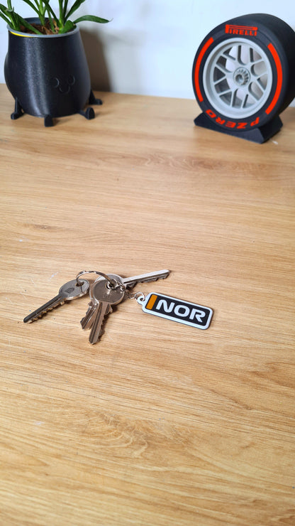Personalized McLaren F1 car keychain with custom "NOR" text on a wooden table, surrounded by other keys, near a whimsical plant pot and a decorative Pirelli tire model.