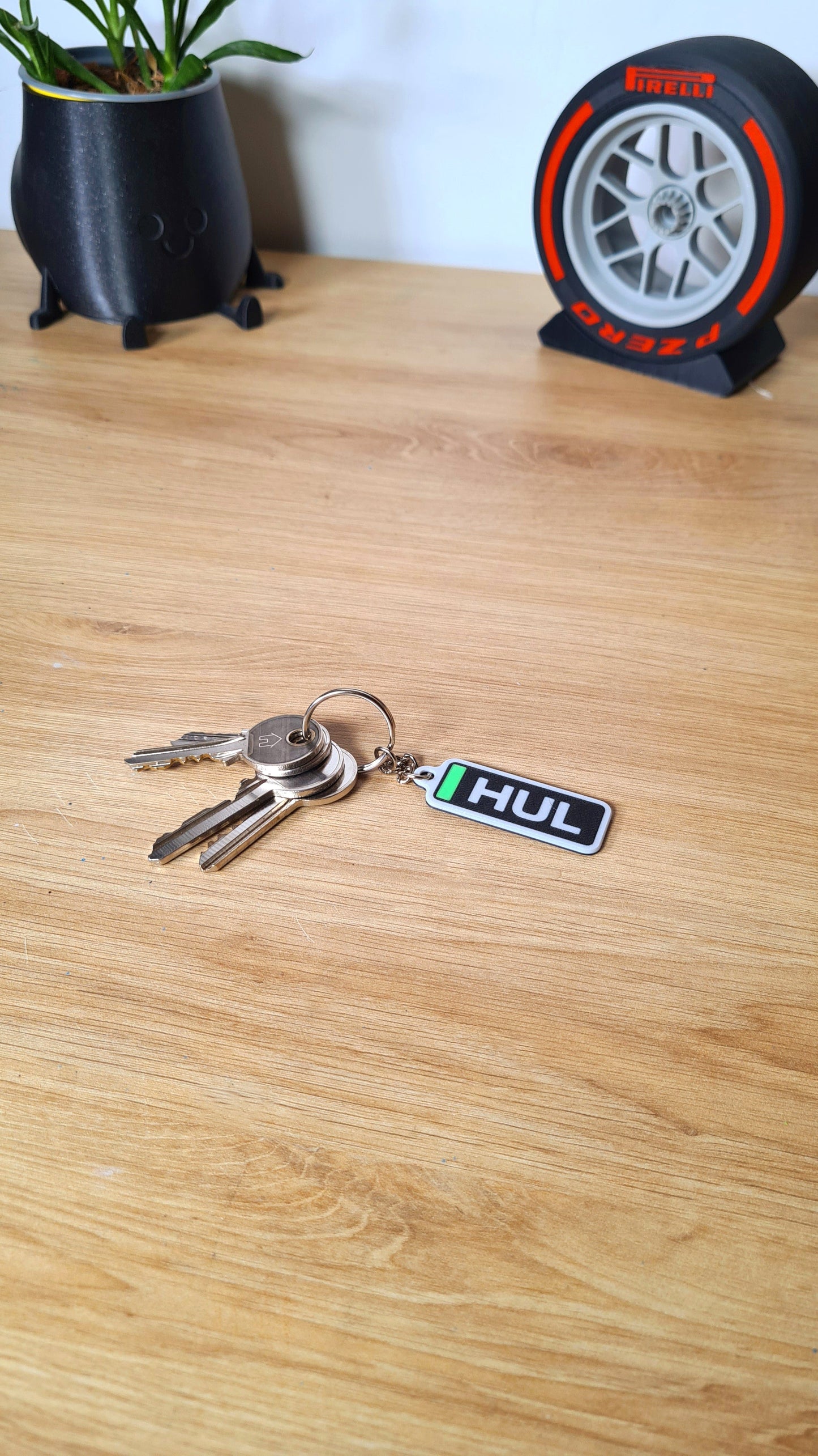 A personalized Formula 1 themed keychain with customizable three-letter tag ("HUL") on a wooden surface next to keys, a smiley plant pot, and a Pirelli tire clock.