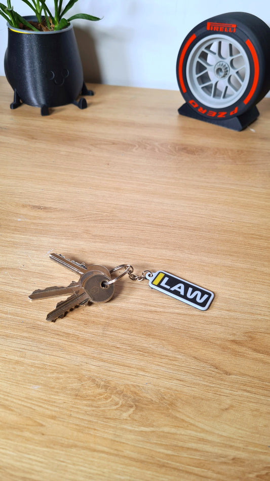 A personalized keychain with the acronym "LAW" in yellow, black, and white on a desk, attached to several keys, near a black plant pot and a Pirelli tire clock.