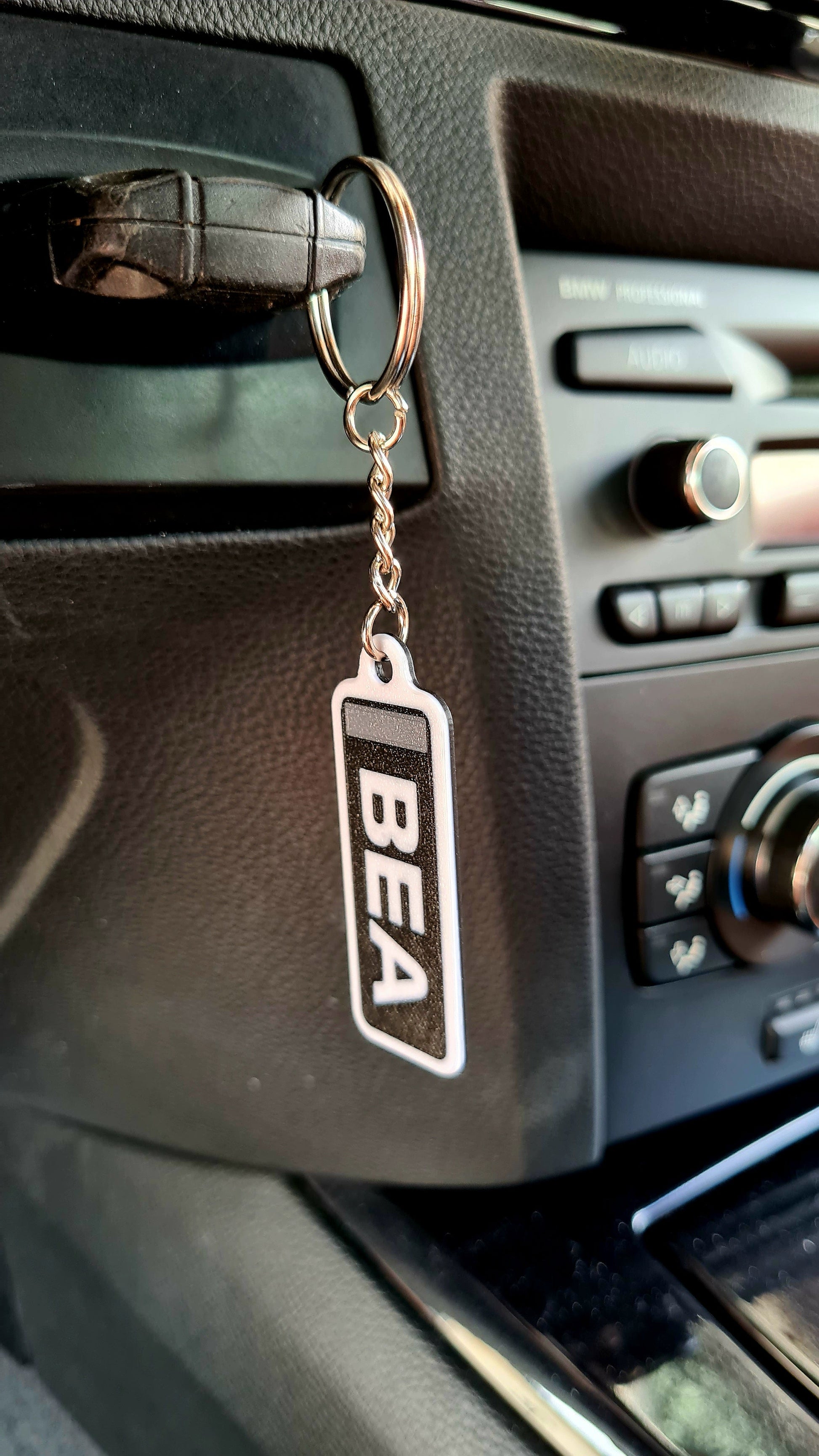 Personalized rectangle keychain with "BEA" in white on a black background attached to a set of three silver keys. Displayed on a wooden table with a car tire clock and a potted plant in the background.