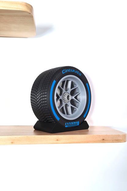 A replica of a Formula 1 wet tire and rim, prominently featuring a detailed tread pattern and blue-accented sidewall, displayed on a black stand against a minimalist background. Perfect for collectors or enthusiasts.