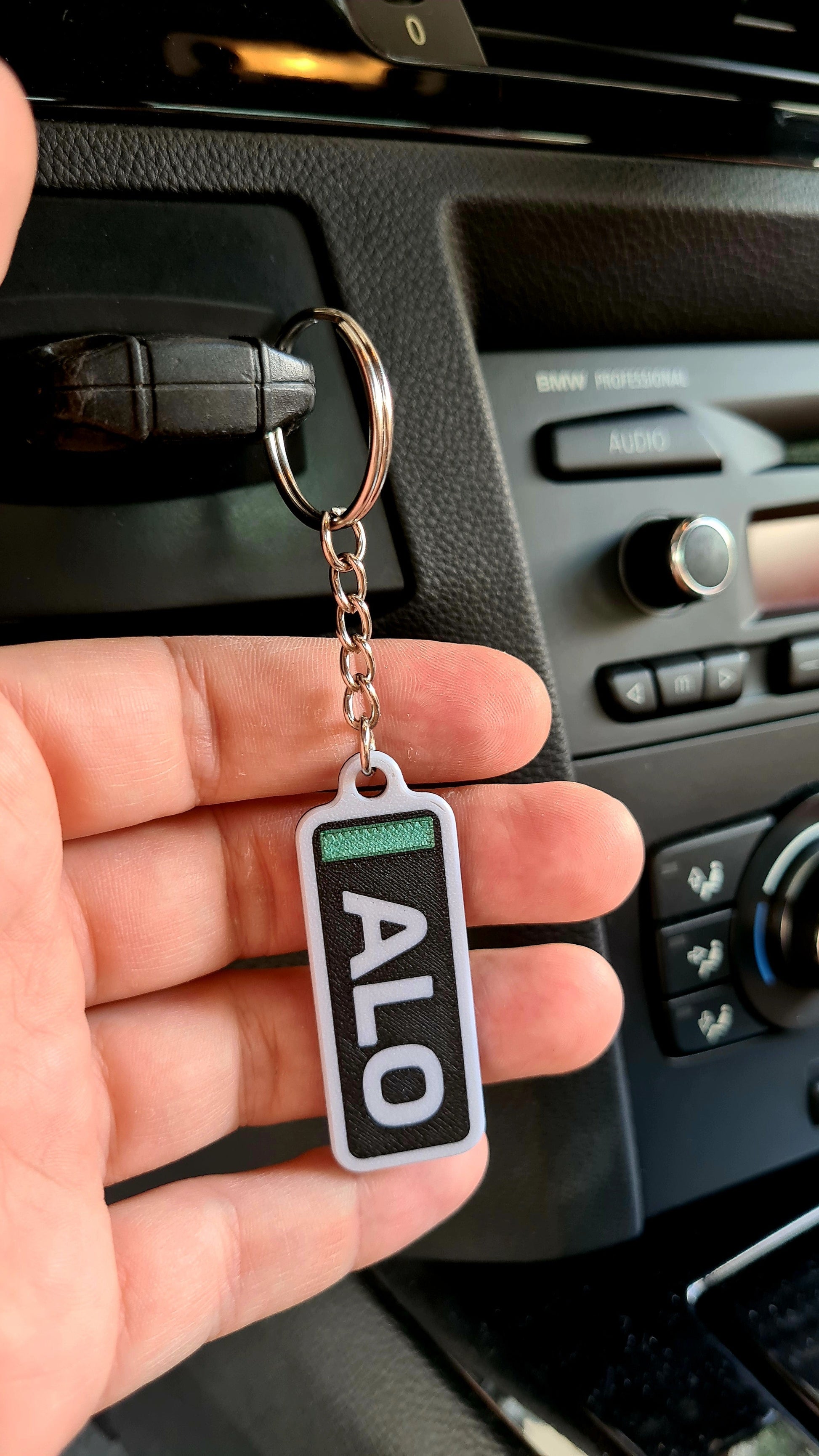 A personalized keychain with the letters "ALO" on a wooden table, alongside house keys, beside a decorative pot and a miniature tire model. Ideal for Formula 1 fans to customize with their favorite driver's initials.