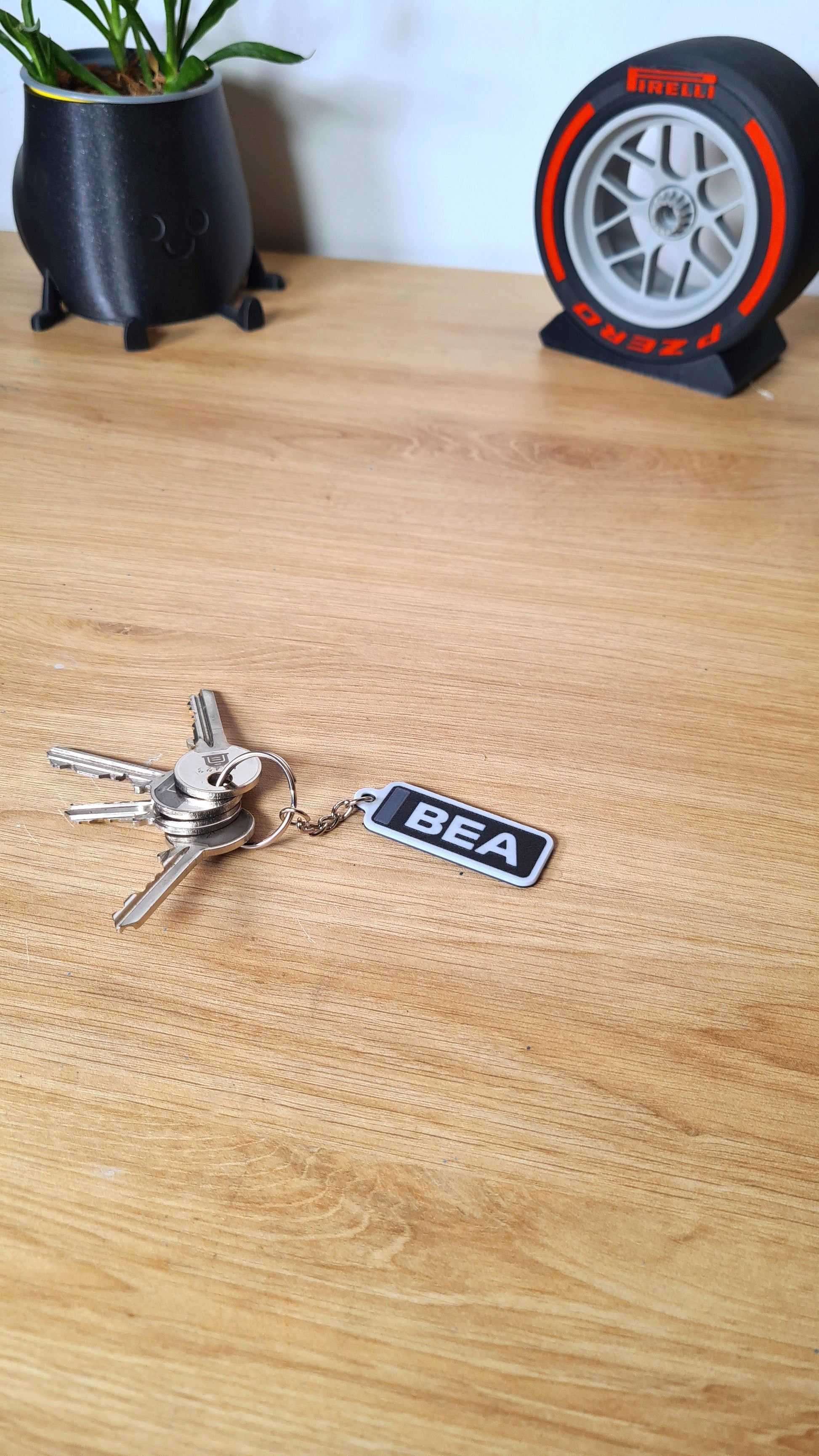 A personalized keychain with the initials "BEA" in white on a black rectangle, attached to a set of keys, lying on a wooden surface next to a potted plant and a Pirelli tire clock.