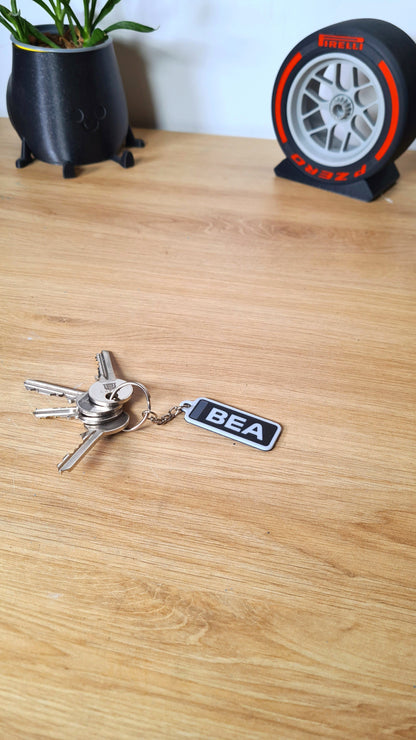 A personalized keychain with the initials "BEA" in white on a black rectangle, attached to a set of keys, lying on a wooden surface next to a potted plant and a Pirelli tire clock.